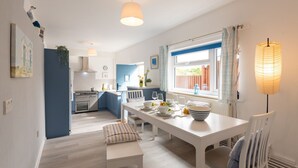 A modern kitchen with white cabinets, a dining area with a white table and chairs, and a window with curtains. - Lan y Môr : Family-Friendly 3‑Bed Cottage with Beach Access in Rhosneigr | Managed by Anglesey Holid (Rhosneigr)