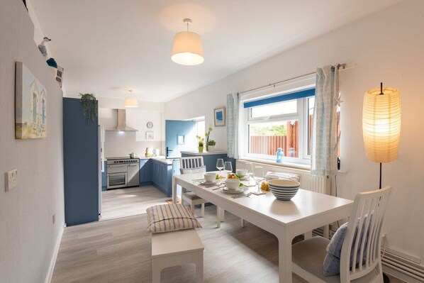 A modern kitchen with a dining area, featuring a white table set for two, a blue accent wall, and a large window with curtains.