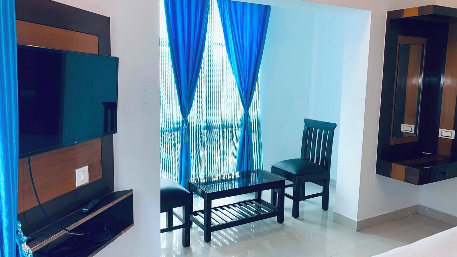 Un salón con televisor de pantalla plana, una mesa de centro de cristal, dos sillones y una ventana con cortinas azules.