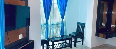 A living room with a flat-screen TV, a glass coffee table, two chairs, and a window with blue curtains.