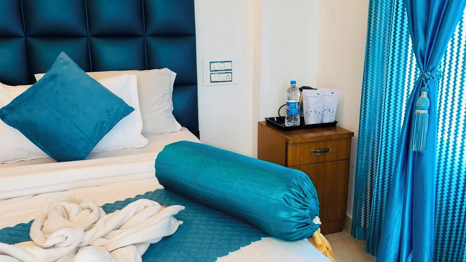 A hotel room with a bed, a wooden nightstand, a water bottle, and a curtain.