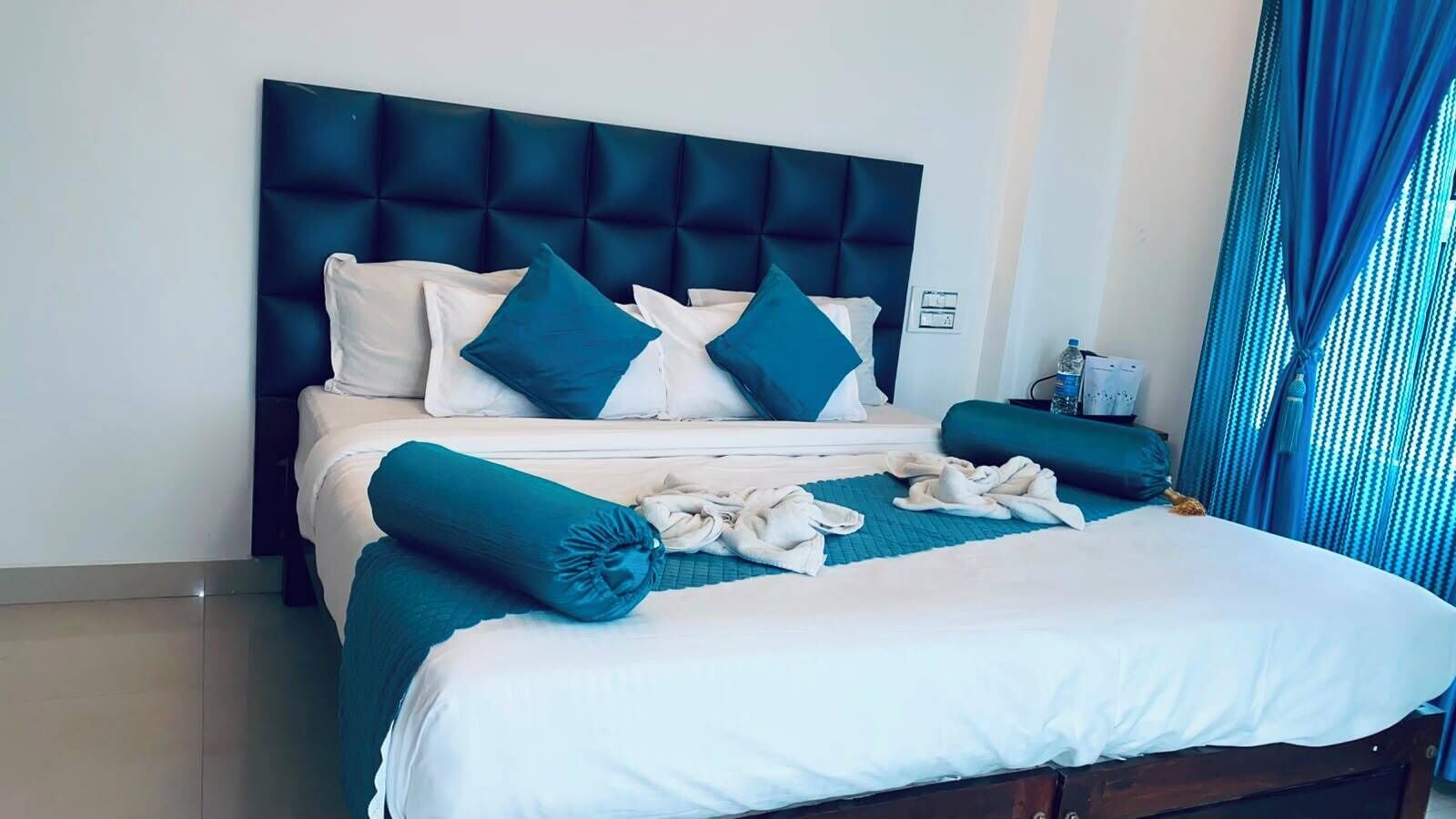 Et hotelværelse med en seng, puder, en sengegavl og et blåt gardin.