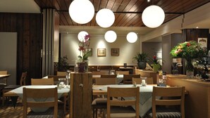 Un interior de restaurante con mesas y sillas de madera, luces esféricas colgantes y plantas en macetas.
