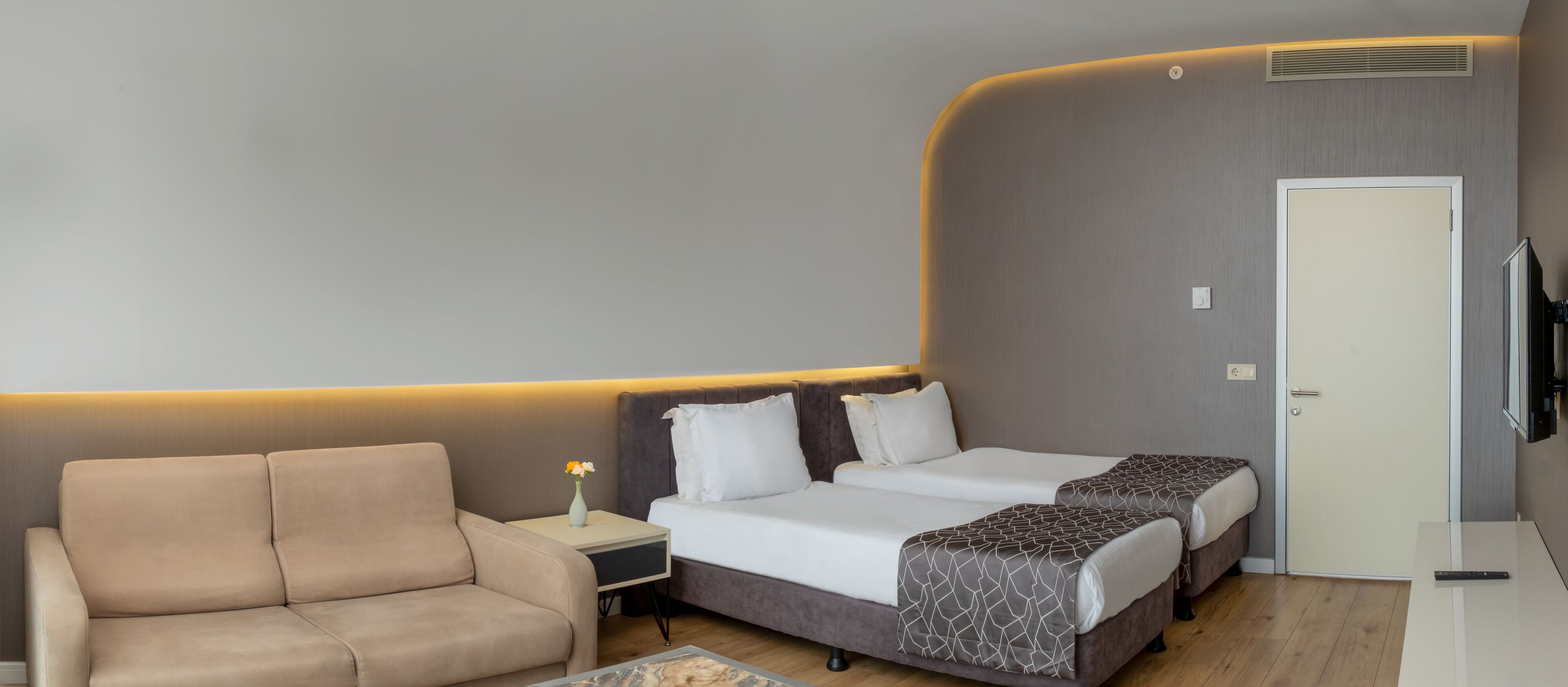 Quarto de hotel com sofá, duas camas, mesa de cabeceira, vaso com flores e televisão.