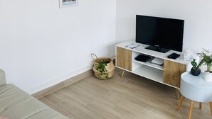 A modern living room with a flat-screen TV on a white cabinet, a beige sofa with a pink pillow, and a small round table with a plant. - Charming Duplex Trouville / Deauville 5mn Train Station (Trouville-sur-Mer)