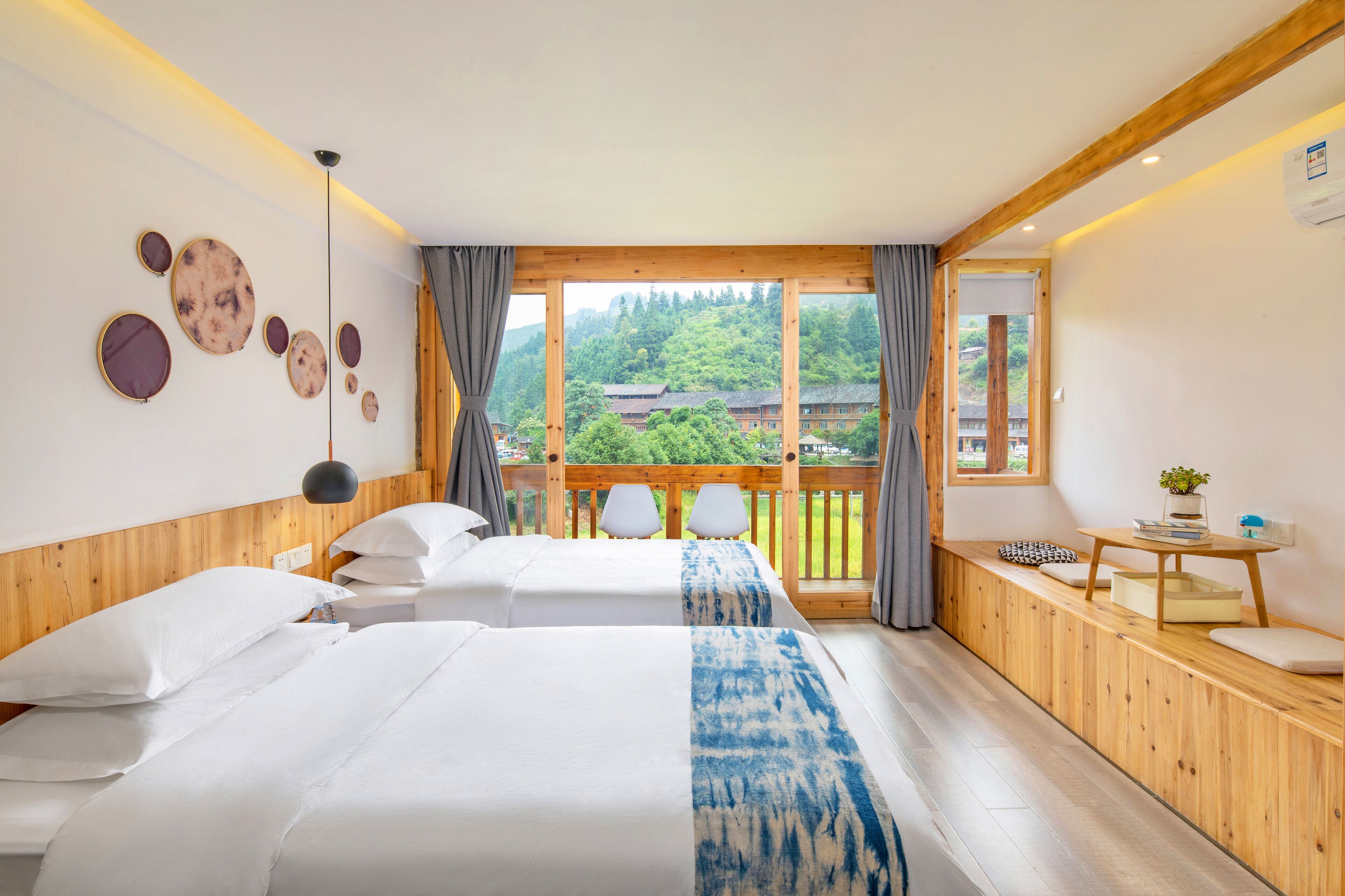 A hotel room with two beds, a wooden bench, a small table with a plant, and a view of a forested area.