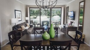 Un coin repas avec une grande table, des chaises et deux vases verts. On y trouve également un canapé et un téléviseur fixé au mur.