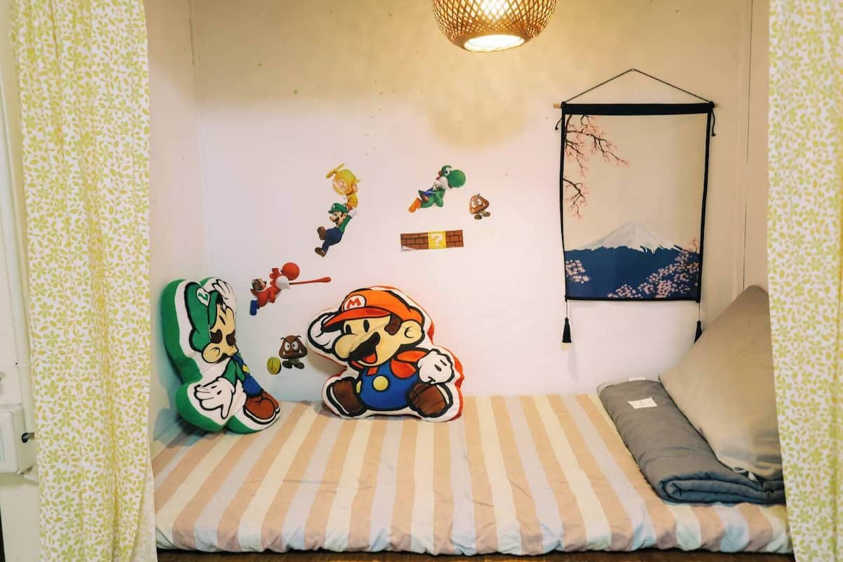 Quarto com cama, almofadas, luminária pendente, decorações nas paredes incluindo um brinquedo de pelúcia do Mario e do Luigi, uma pequena pintura e uma prateleira com alguns objetos.
