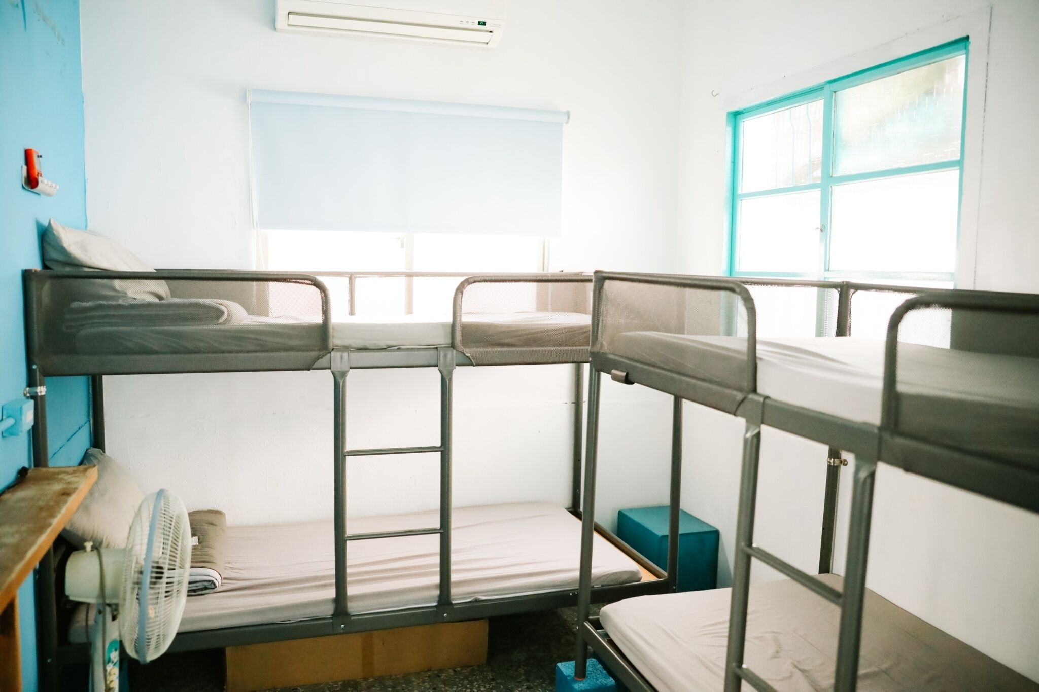 Un dormitorio con literas, tres literas, una ventana con persiana azul, un ventilador y una pequeña caja azul en el suelo.