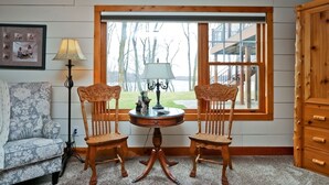 Een gezellige kamer met een houten tafel en twee stoelen, een lamp, een bank en een raam met uitzicht op bomen en gebouwen.
