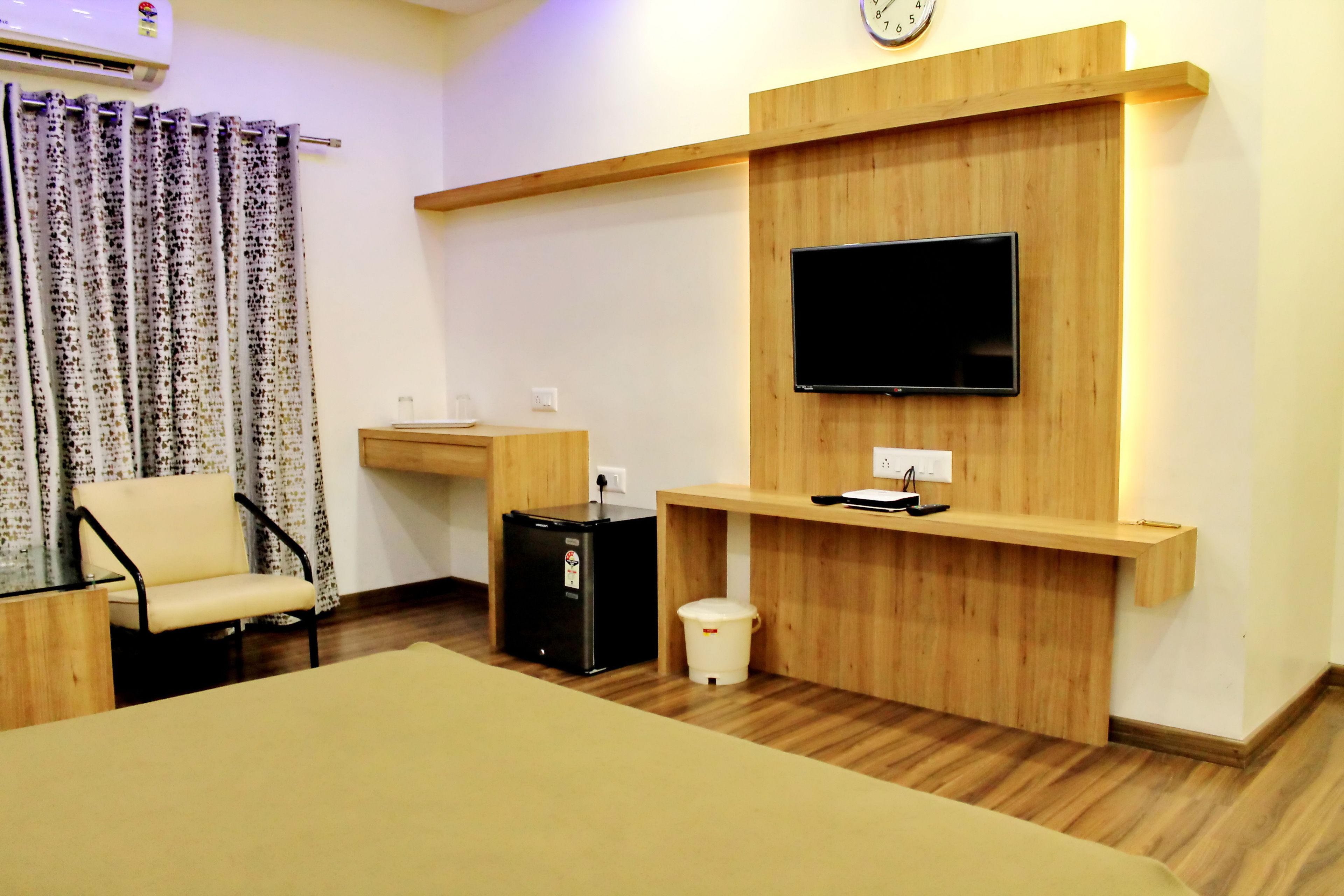 A hotel room with a wooden wall unit, a flat-screen TV, a small desk, a chair, a black refrigerator, and a trash bin.