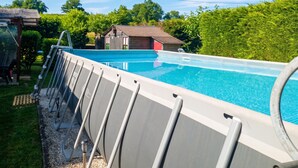 A swimming pool with a metal deck, surrounded by greenery and a small building.