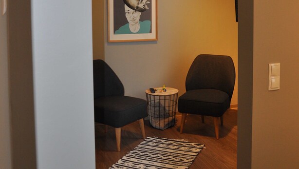 A hallway with two black chairs, a small round table, a framed picture on the wall, and a patterned rug on the floor. - Gästehaus Sonne (Triefenstein)