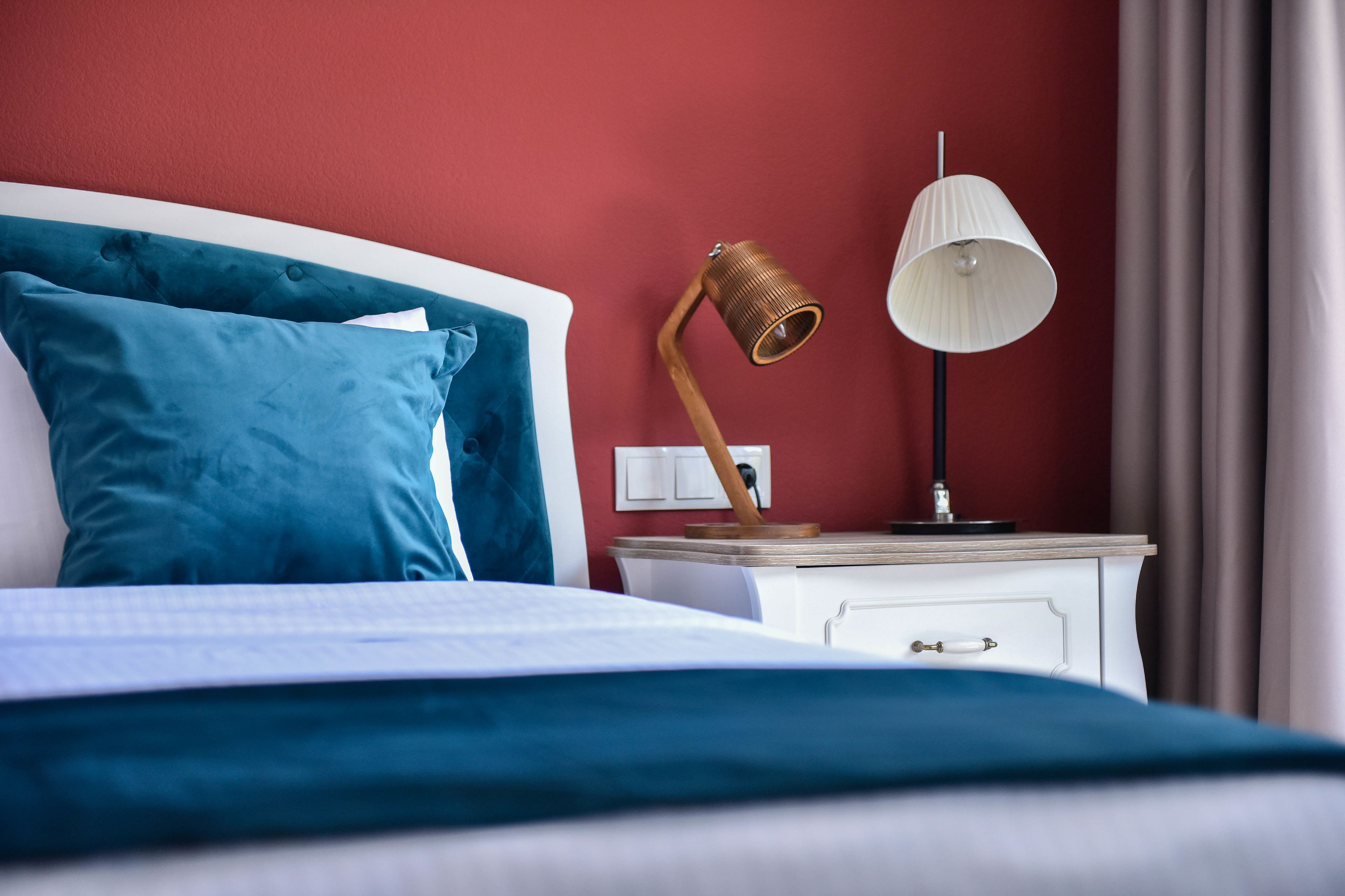 A neatly made bed with blue bedding, a white bedside table with a lamp, and a red wall.