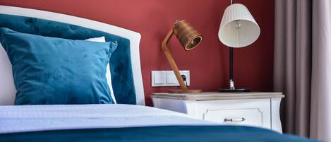 A neatly made bed with blue bedding, a white bedside table with a lamp, and a red wall.