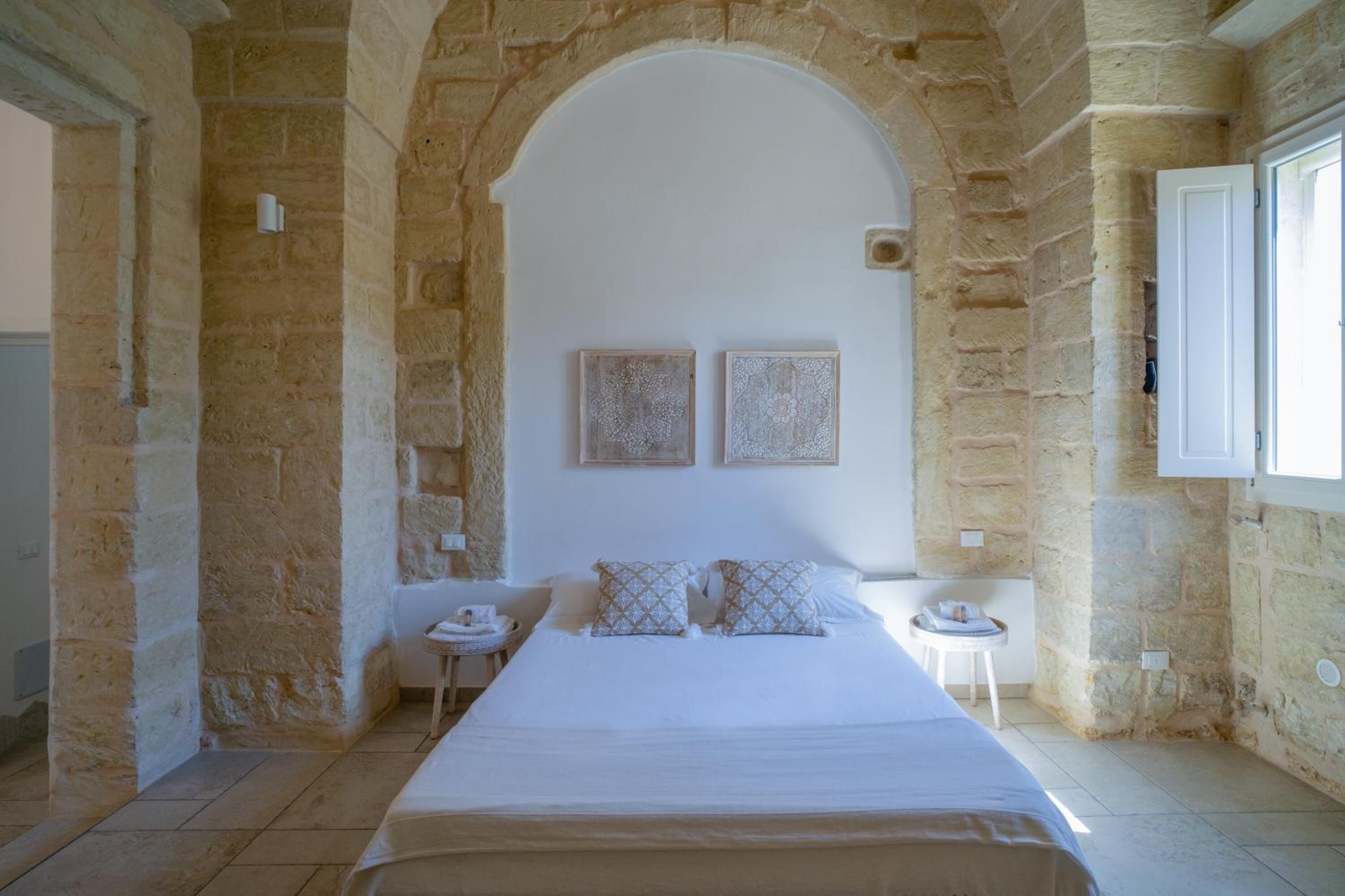A bedroom with a stone archway, a bed with white linens, two bedside tables, and a window with a white blind.