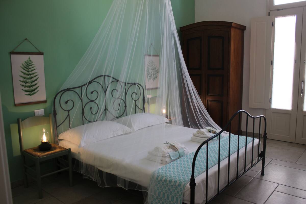 Un lit bien fait, surmonté d’un dais, une table de chevet avec une lampe et une plante encadrée accrochée au mur.