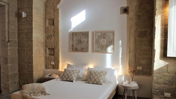 A bedroom with stone walls, a bed with white linens and patterned pillows, a bedside table with a lamp, and two wall-mounted decorative pieces. - Li Perrini Boutique B&B (Vernole)