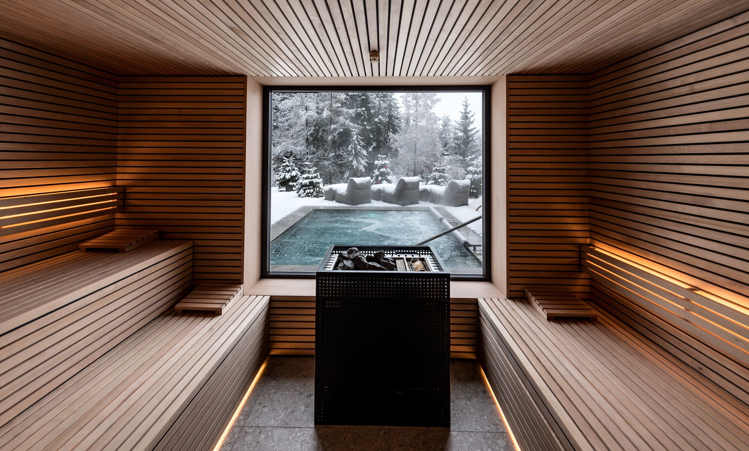 Una sauna con listones de madera, un banco y un ventanal con vistas a un bosque nevado.