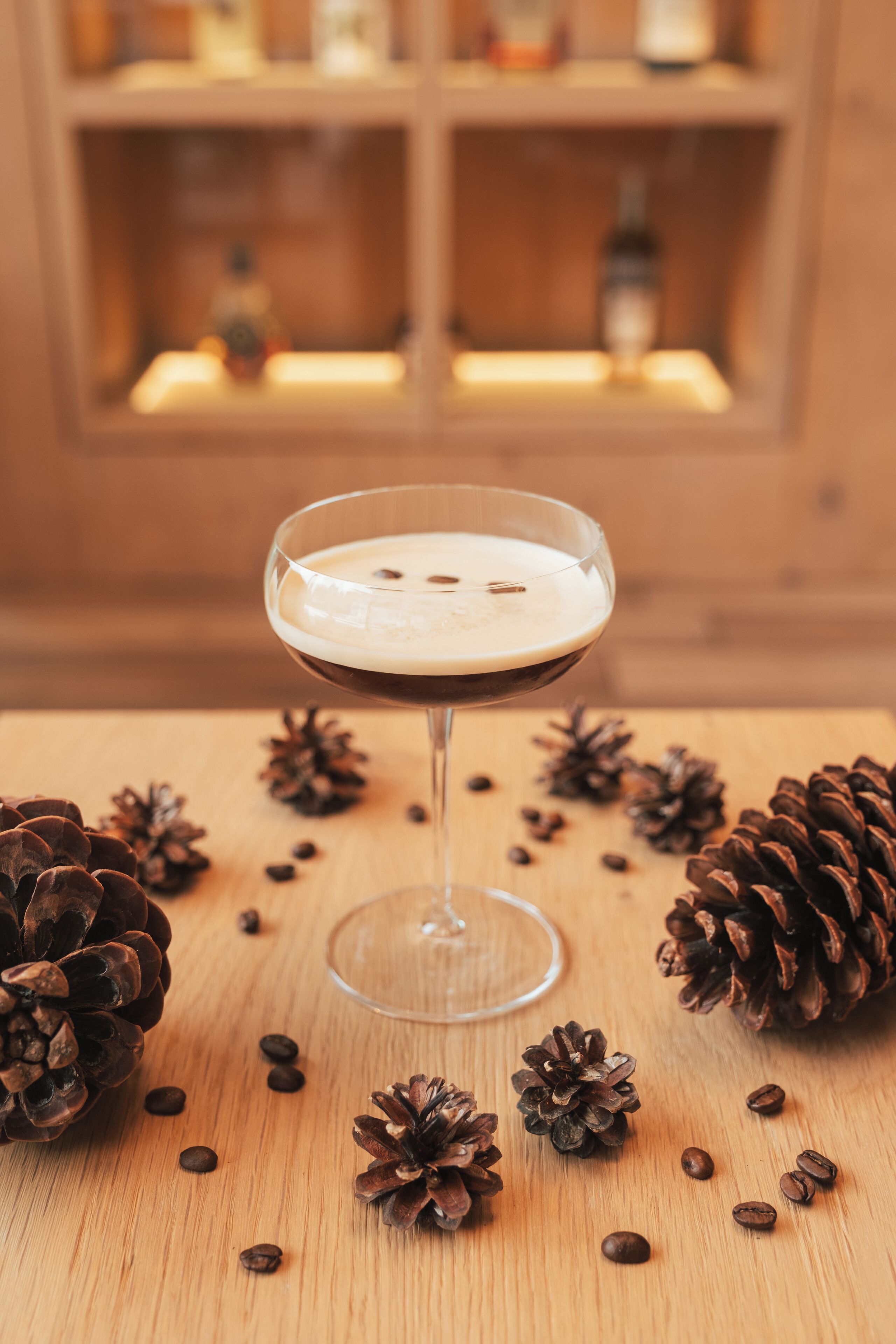 A cocktail glass with a frothy drink on a wooden surface surrounded by pine cones and coffee beans.
