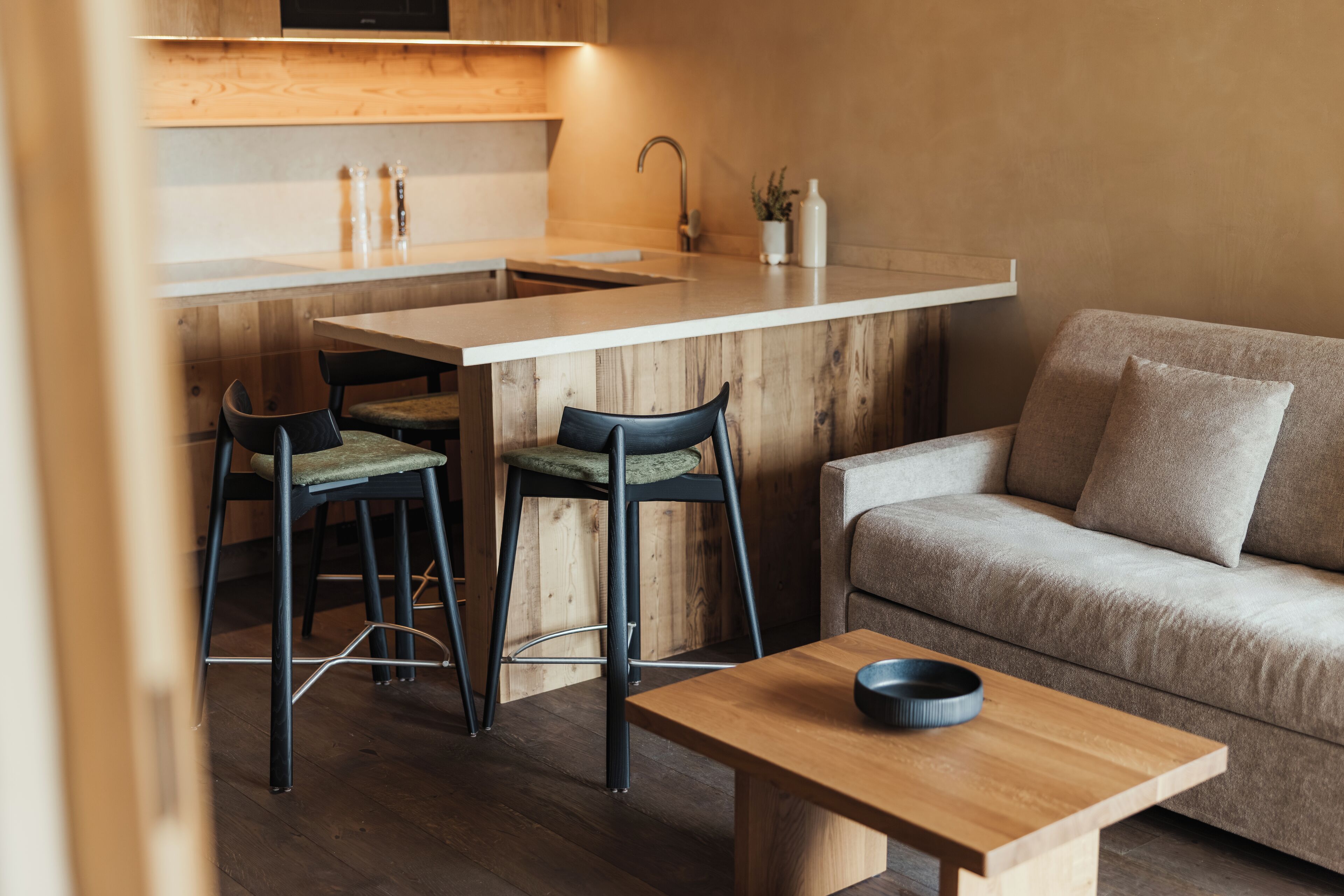 A compact kitchen with a wooden countertop, two bar stools, a dining table, and a sofa.