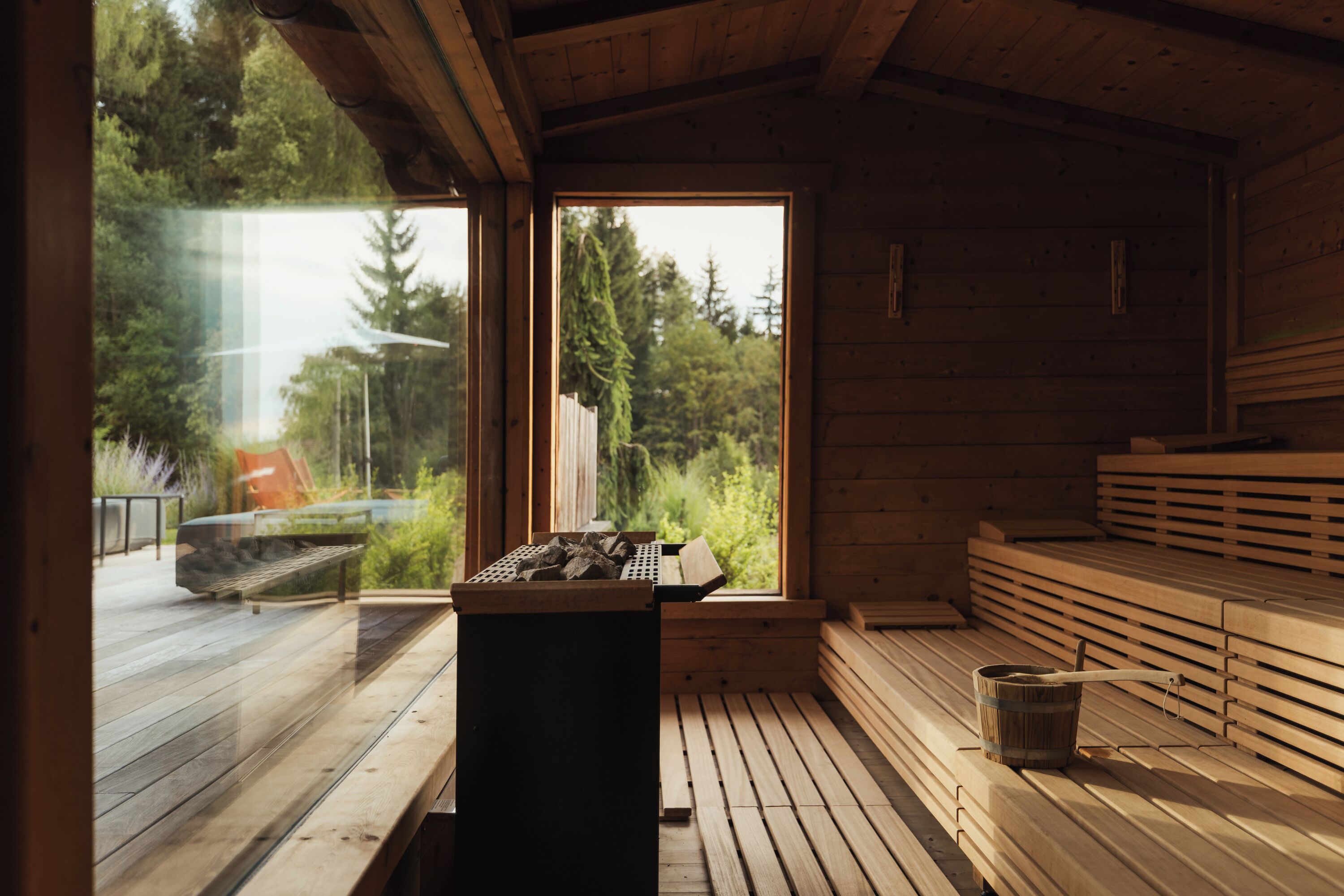 A wooden sauna with benches and a sauna table, large windows offering a view of trees and a deck outside.