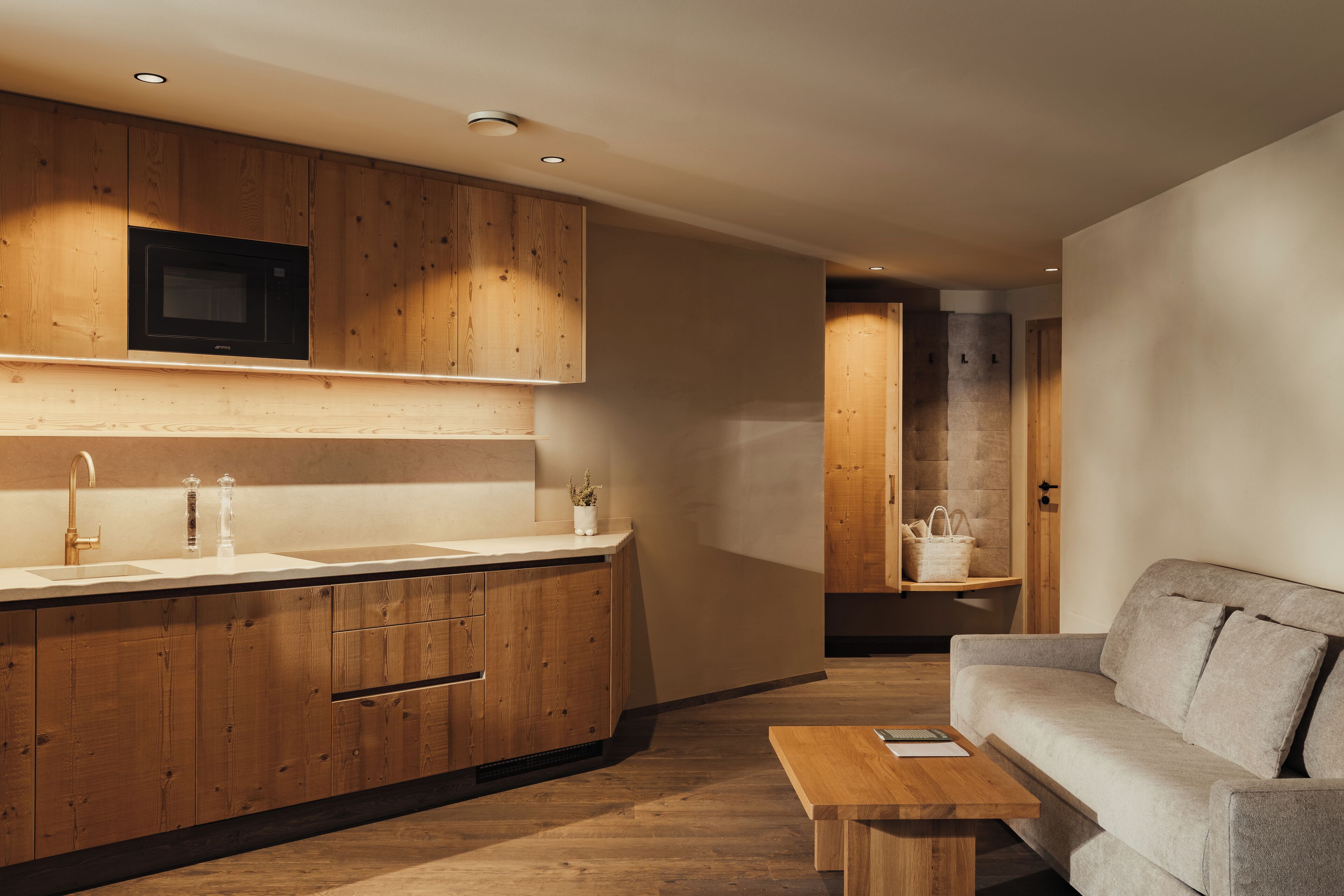 Una cocina moderna con armarios de madera, un microondas y un lavamanos. En la sala de estar, hay un banco de madera y un sofá gris.