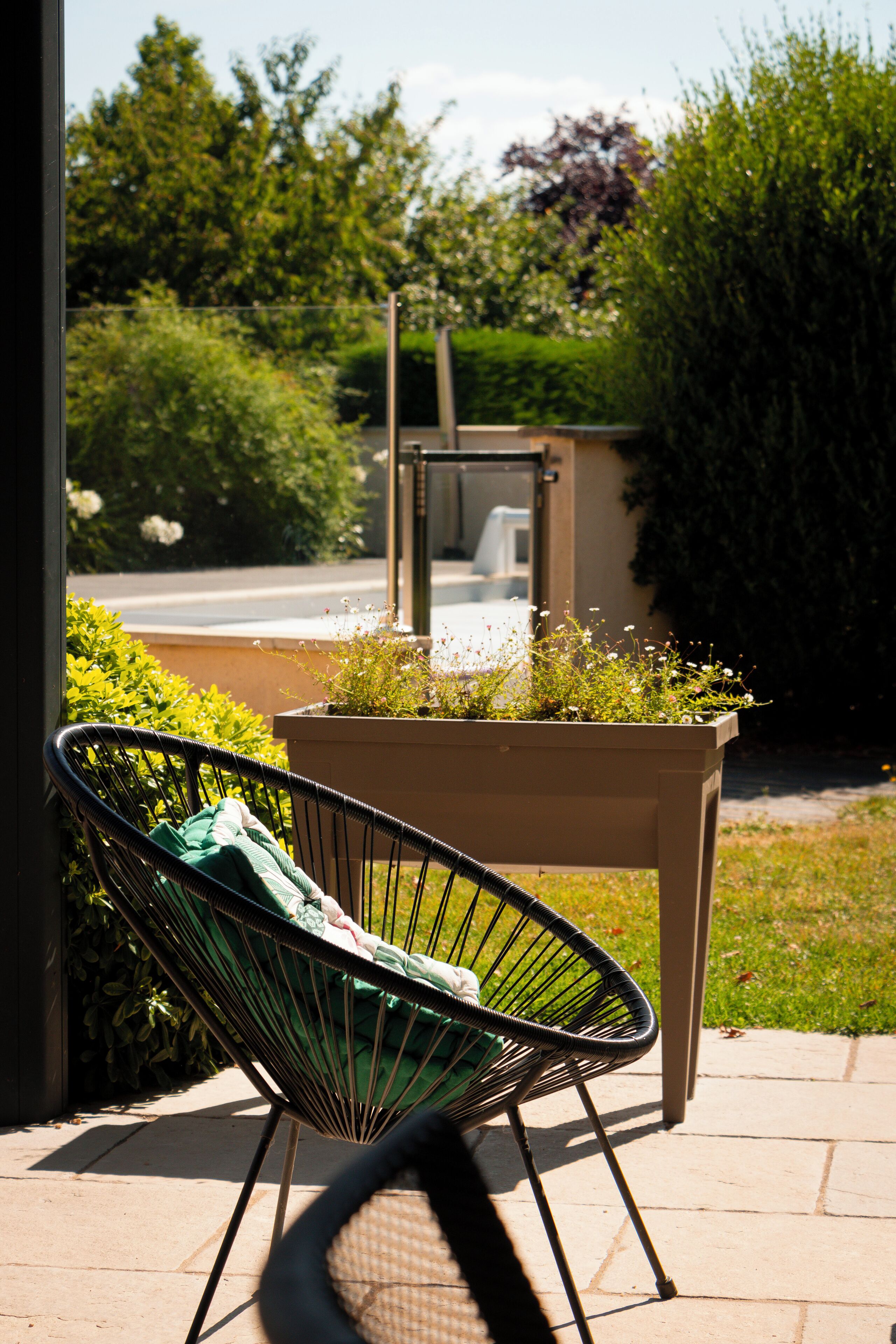 A wicker chair with a cushion, a planter box with greenery, and a small structure in the background.