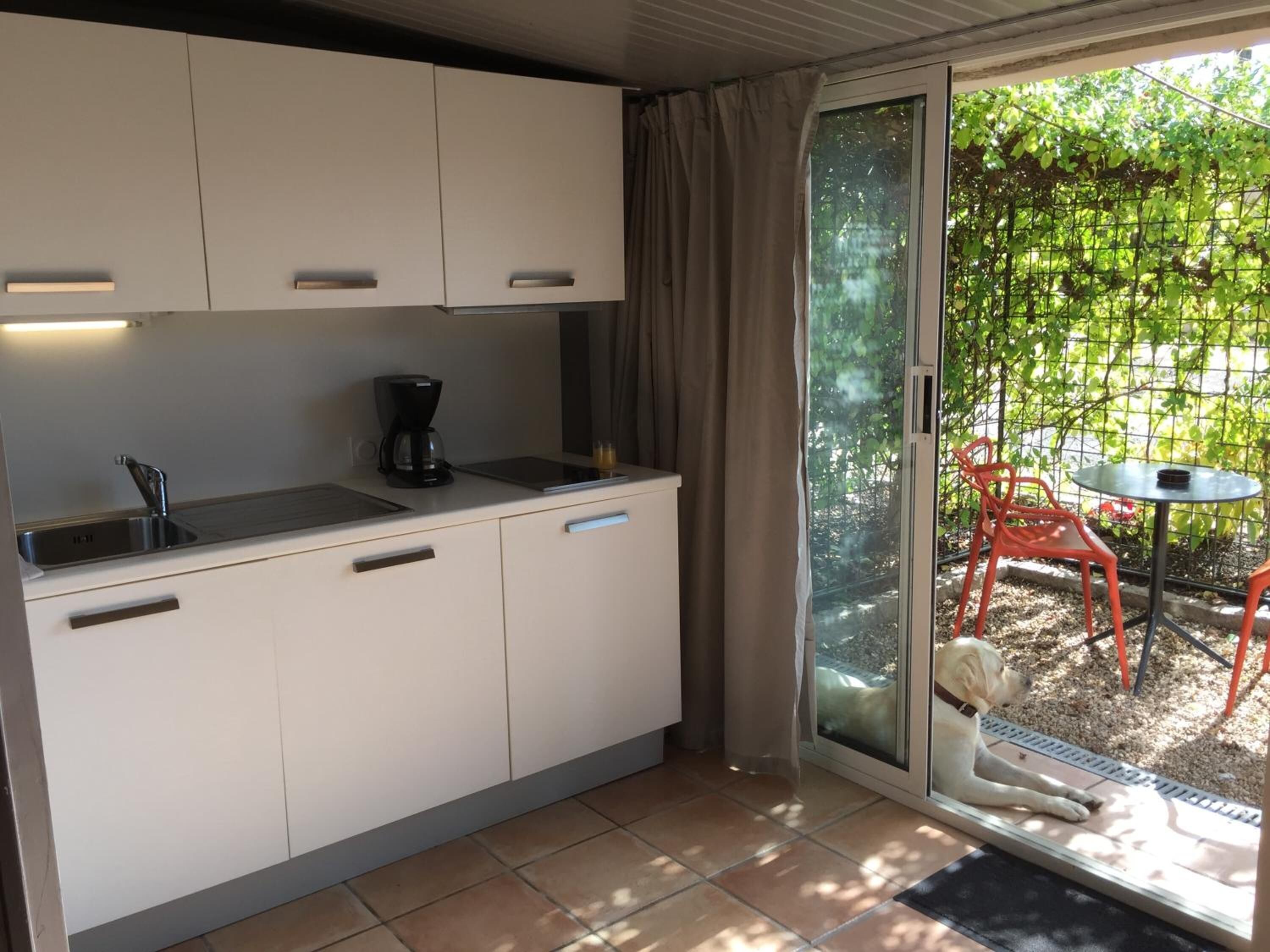 A compact kitchen with a sink, a coffee maker, and cabinets. There is a sliding glass door leading to an outdoor area with a table and chairs.