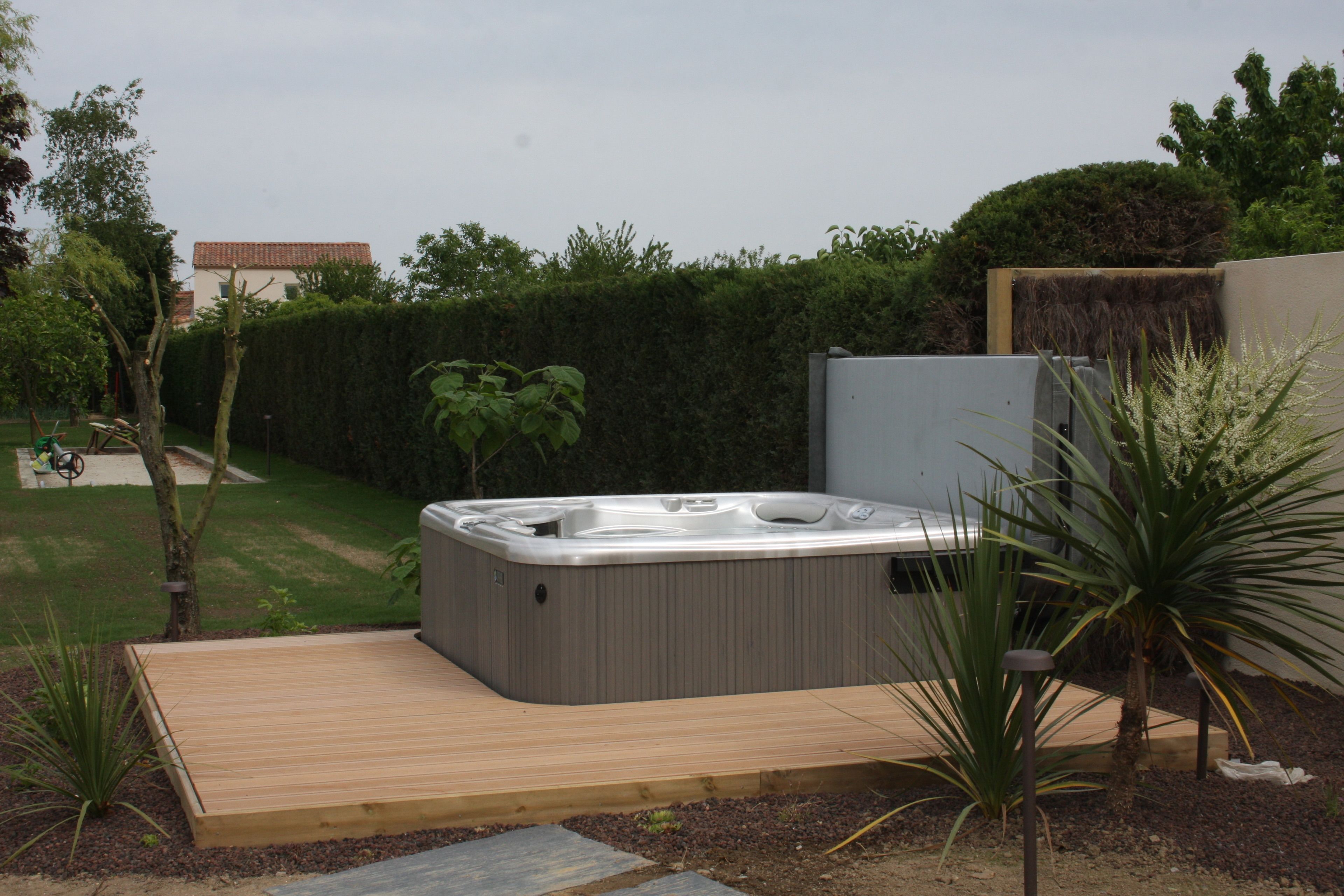 A hot tub in a garden with a wooden deck, surrounded by plants and a hedge.