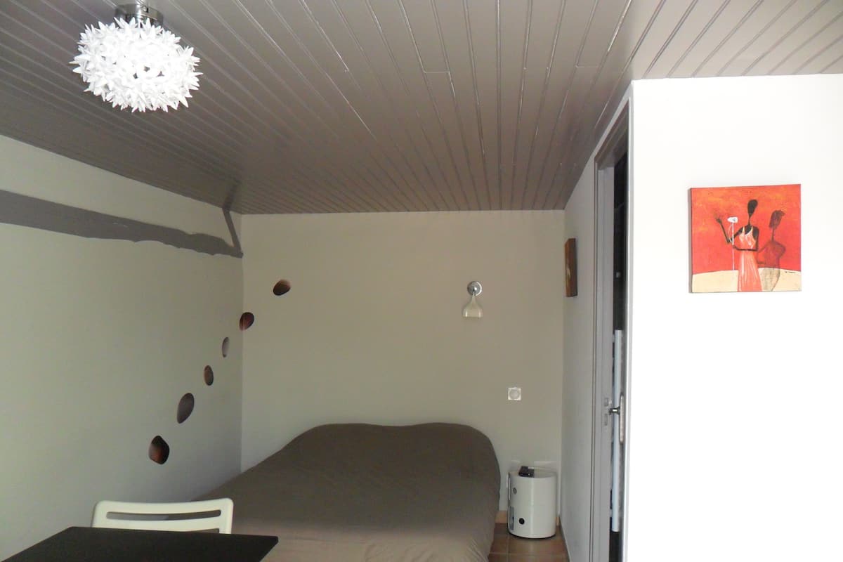 Une chambre à coucher avec un lit, une chaise blanche, une petite table, une lampe blanche, une poubelle et un tableau accroché au mur.