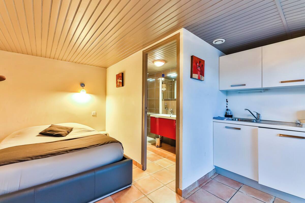 Une chambre à coucher avec un lit, une lampe de chevet et une vue sur une cuisine dotée de meubles blancs et d’un plan de travail rouge.