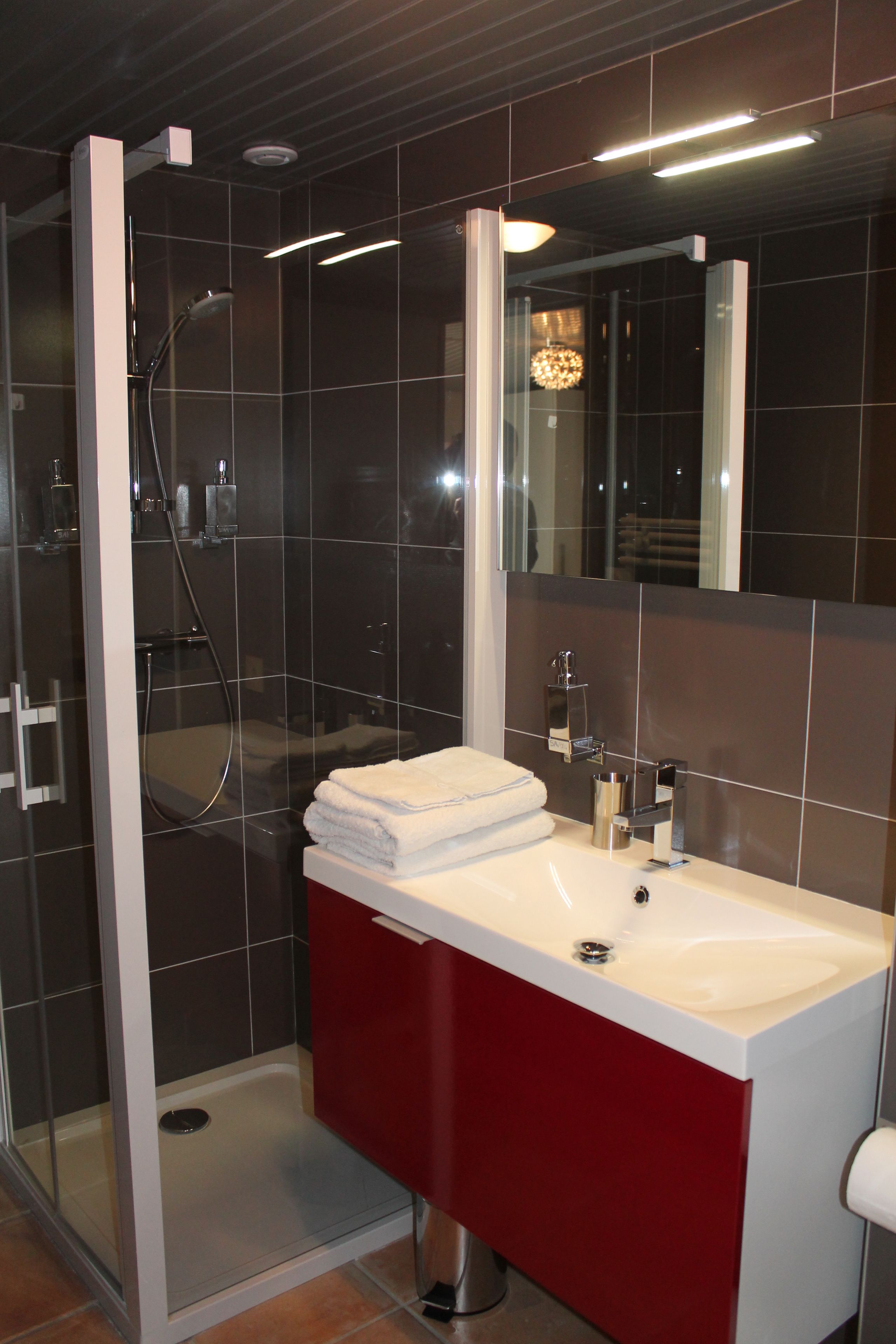 A modern bathroom with a glass-enclosed shower, a white sink with a red cabinet, and a large mirror.