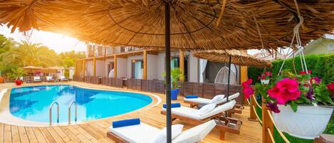 Um deck de madeira com piscina, cadeiras de descanso e um guarda-sol de palha.
