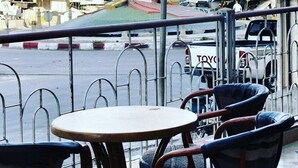 A view from a balcony with a table and chairs, overlooking a street with parked cars and a red structure in the background. - Hotel El Kssour (Medenine)