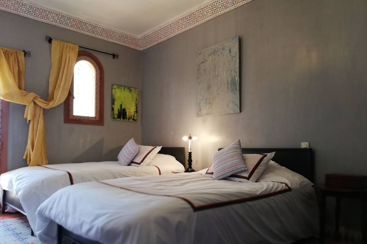 Una cama doble con ropa de cama blanca y almohadas a rayas, una mesita de noche con lámpara y una ventana con cortinas amarillas.