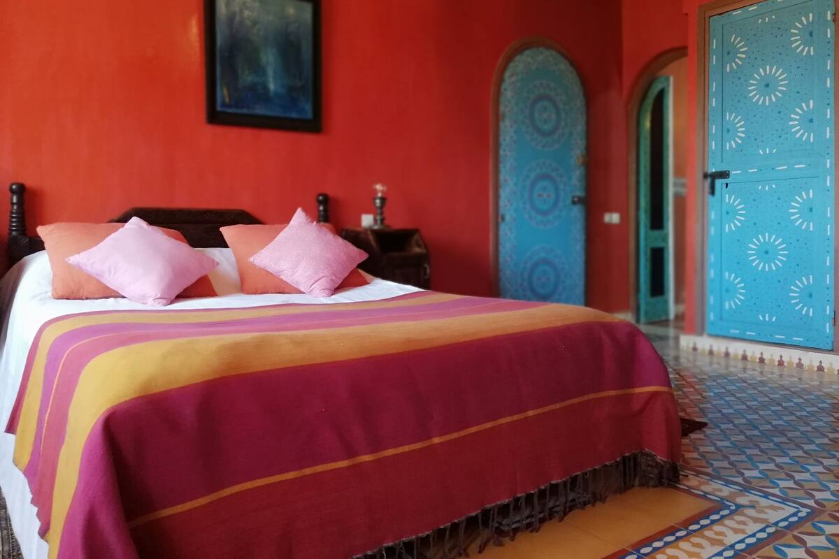 Una cama con una colcha a rayas de colores y varias almohadas, un cabecero de madera oscura, un cuadro en la pared y una puerta azul con diseños decorativos.