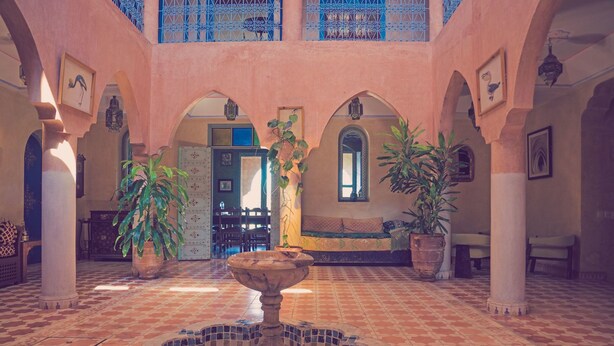 A courtyard with a fountain, arches, potted plants, and a tiled floor.