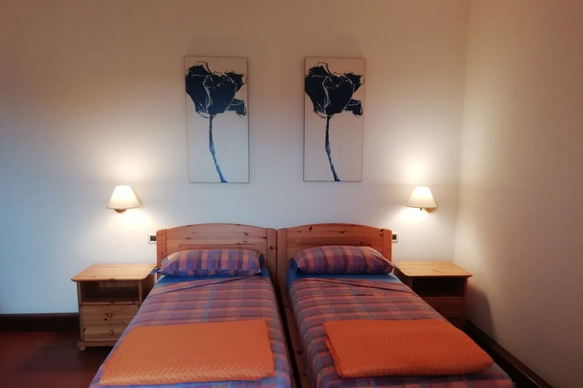 A double room with two beds, each with a plaid duvet and an orange blanket. There are two wall-mounted lamps on either side of the beds.