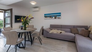 A modern living room with a grey sofa, a round dining table with chairs, a TV mounted on the wall, and a painting of a beach scene. - Villa Irma in Golas, Croatia (Golas)