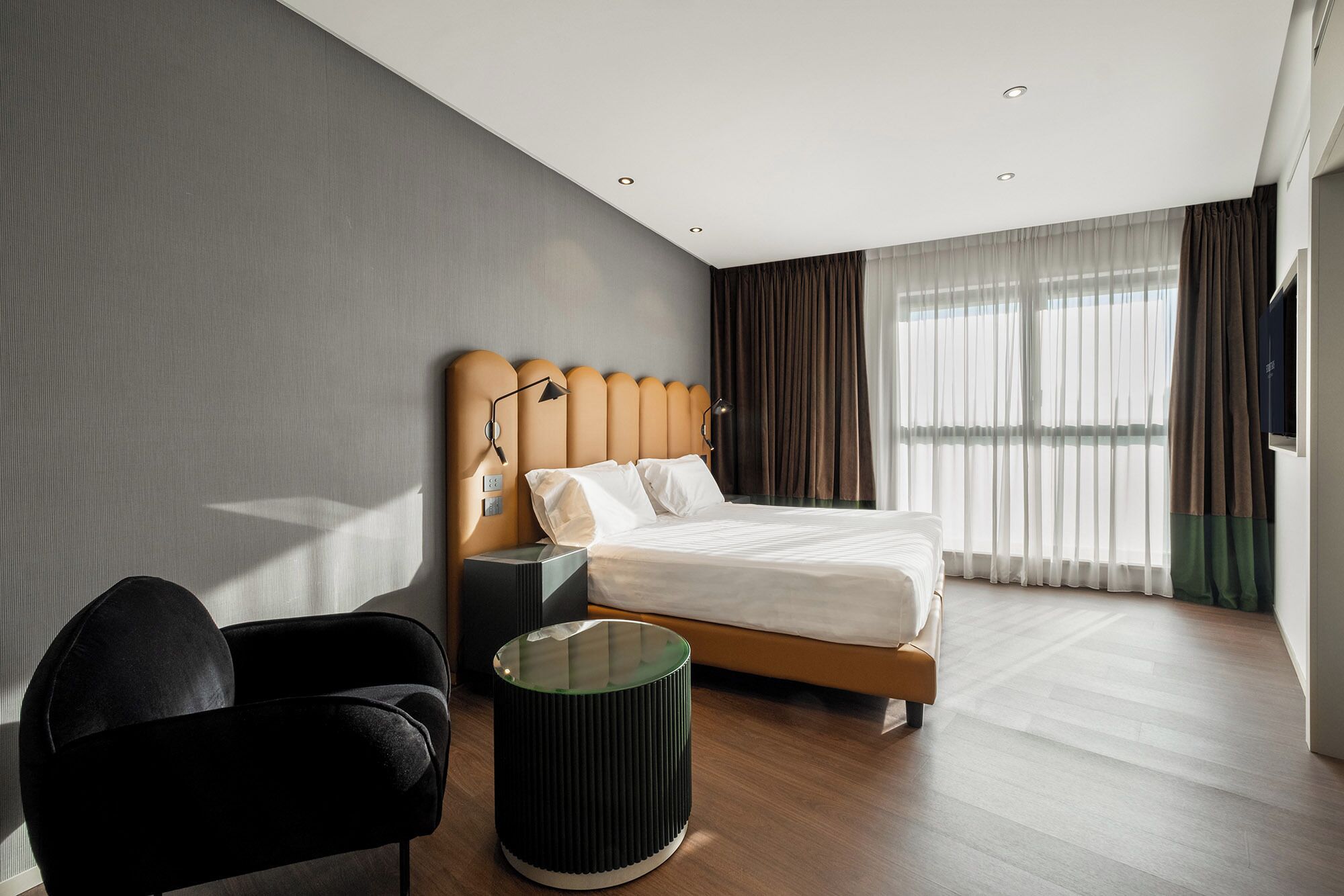 Una habitación de hotel moderna con una cama grande, una mesita auxiliar, una silla y una ventana con cortinas.