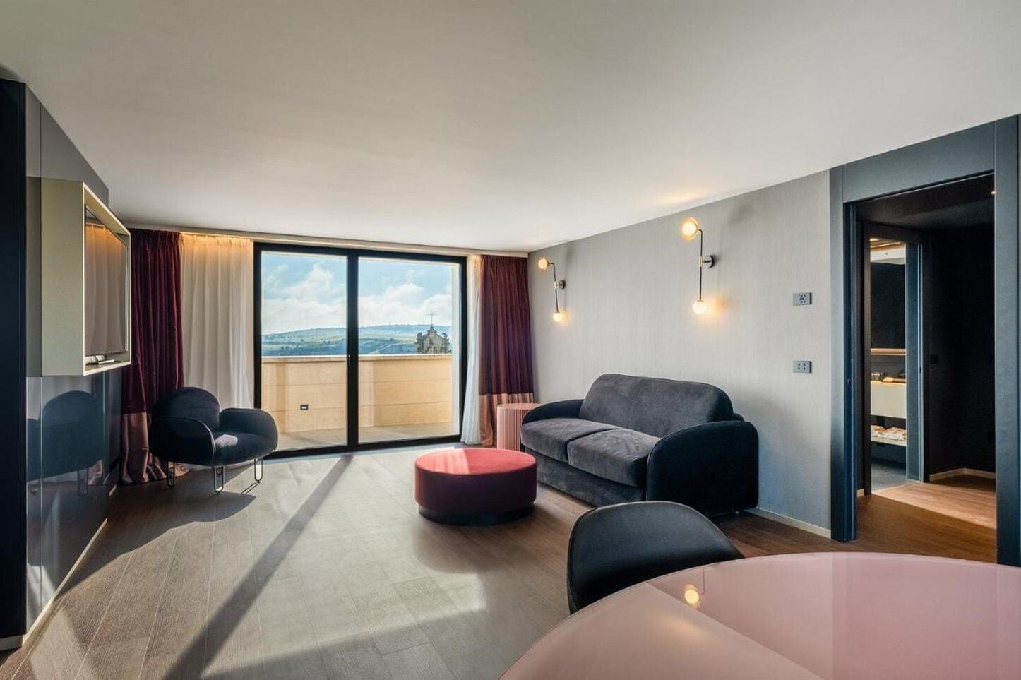 A modern living room with a sofa, a round ottoman, and a dining table. There is a large sliding glass door leading to a balcony with a view of the countryside.