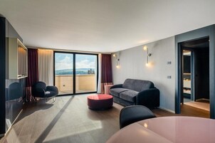 A modern living room with a sofa, a round ottoman, and a dining table. There is a large sliding glass door leading to a balcony with a view of the countryside.