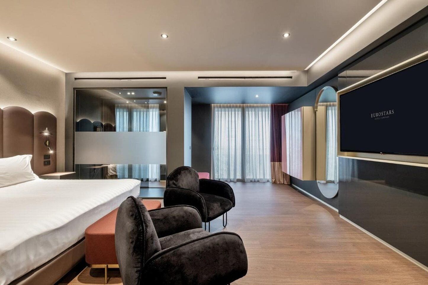 Una moderna camera d'albergo con un letto grande, un'area salotto con due poltrone, una TV a schermo piatto e un ampio specchio.