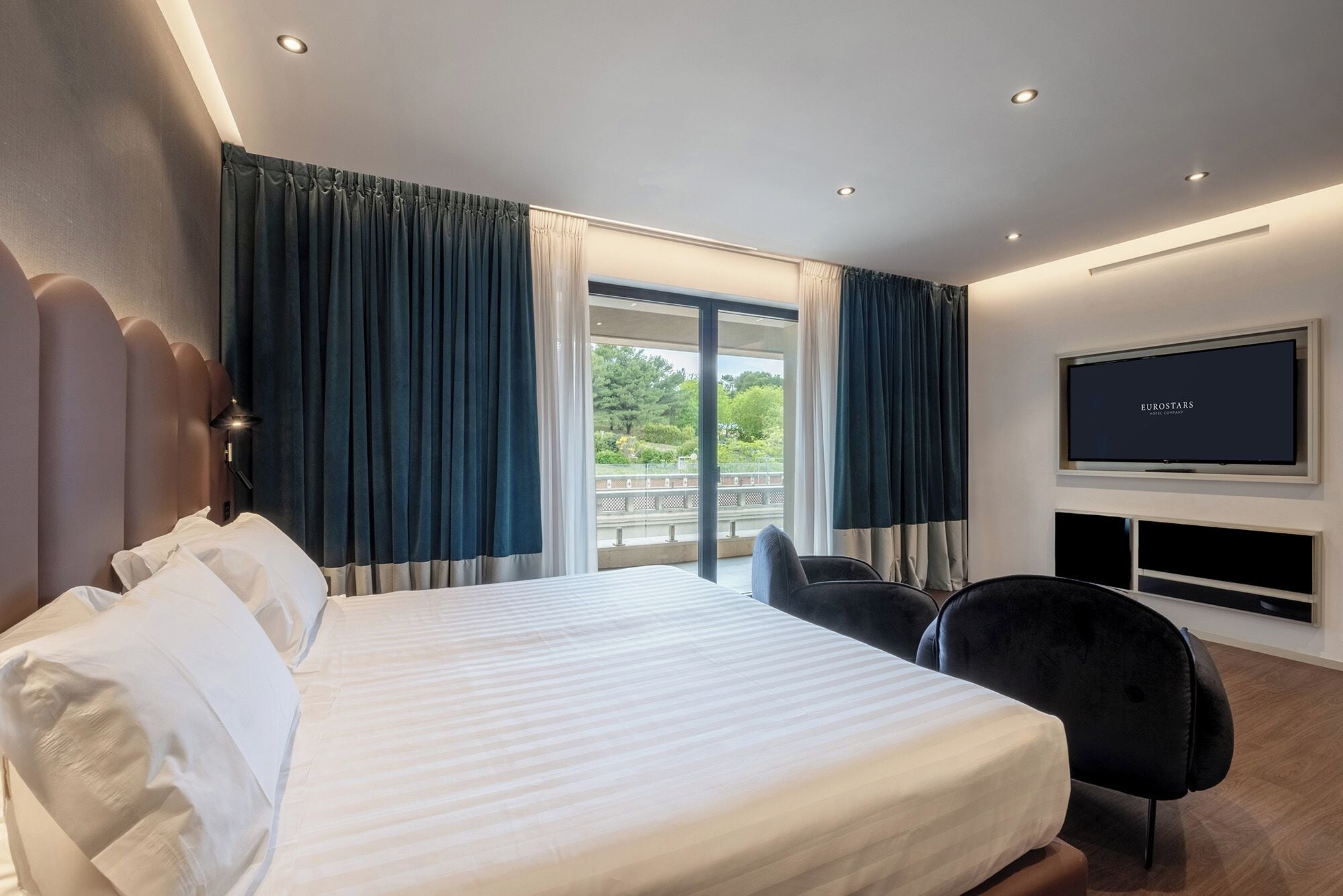 A hotel room with a large bed, a TV, and a balcony with curtains.
