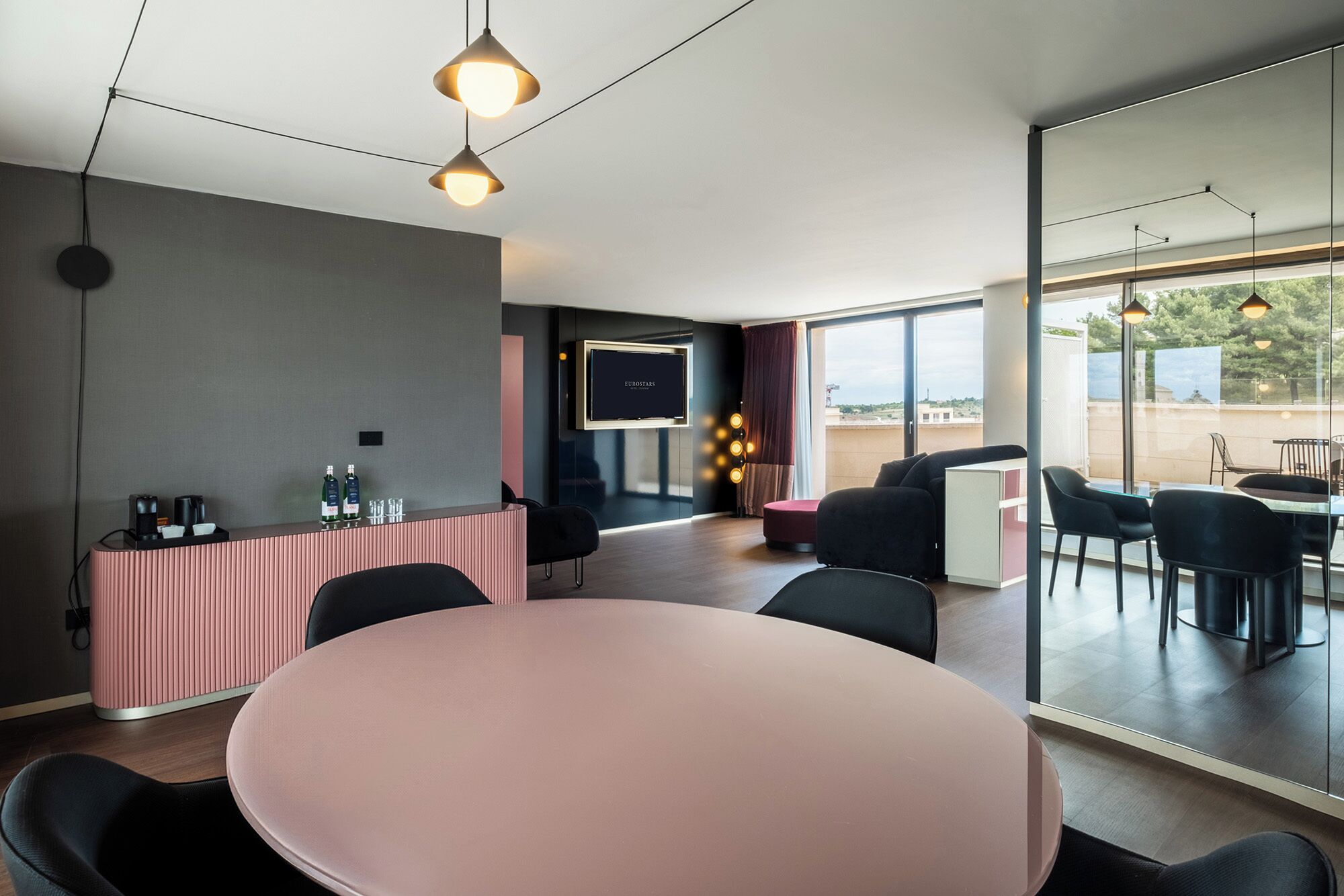 Habitació d'hotel moderna amb una taula rodona, cadires negres, un taulell de recepció rosa, un televisor de pantalla plana i una finestra gran que ofereix vistes a l'exterior.