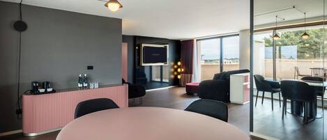 Habitació d'hotel moderna amb una taula rodona, cadires negres, un taulell de recepció rosa, un televisor de pantalla plana i una finestra gran que ofereix vistes a l'exterior.