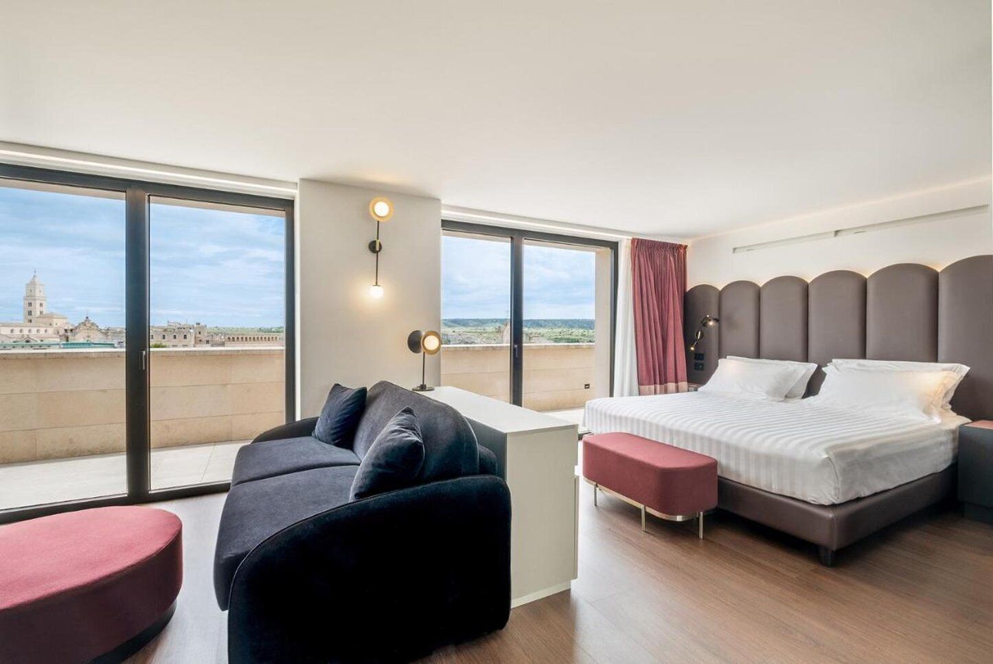 Una camera d'albergo moderna con un letto grande, un divano, un tavolino e vista sulla città attraverso ampie finestre.