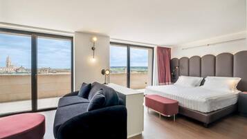 Una camera d'albergo moderna con un letto grande, un divano, un tavolino e vista sulla città attraverso ampie finestre.
