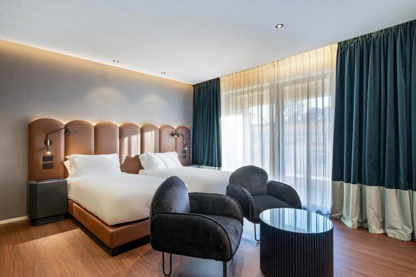 A modern hotel room with a large bed, two armchairs, a small round table, and a nightstand with a lamp.